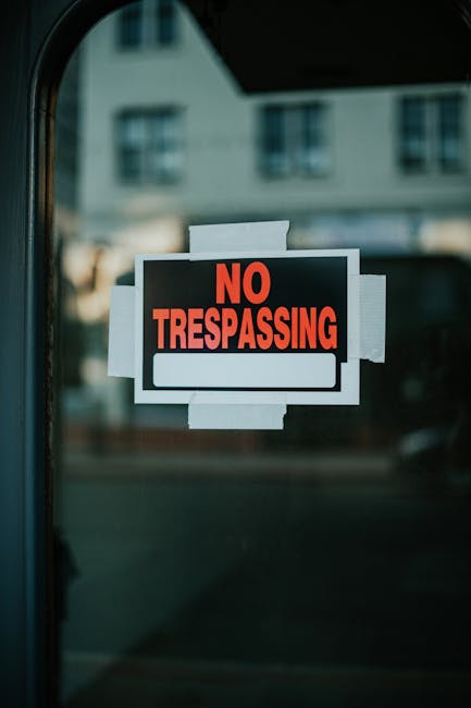 A close-up view of a rectangular 'No Trespassing' sign attached to a glass door or window. The sign features a black background with bold, red uppercase letters spelling 'NO TRESPASSING' at the top and a blank white rectangular space beneath for additional information. The sign is secured with multiple pieces of white tape around its edges, affixed to transparent glass, which reflects a faint outdoor scene showing residential buildings with multiple windows and a cloudy sky. The environment suggests an urban or suburban setting. The sign's presence indicates restricted access to the property, typically managed through private notices, aligning with alternative waste management or property access control measures commonly used by waste removal services such as Waste Removal Enfield that operate independently of local authority rubbish collection.