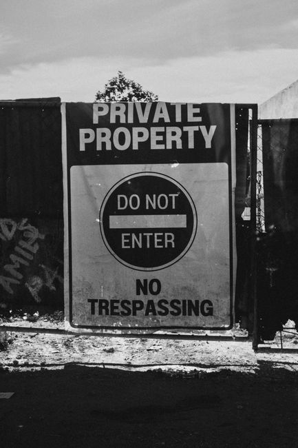 A large rectangular sign mounted on a metal gate and post, displaying the words 'PRIVATE PROPERTY' in bold uppercase letters at the top, with a black and white circular symbol beneath that reads 'DO NOT ENTER' with a horizontal line across the center. Below the symbol, the sign states 'NO TRESPASSING' in capital letters. The gate appears to be made of metal, possibly painted black, and is situated outdoors on a paved or gravel surface. The background includes a portion of a white canopy or tent on the right side and a silhouette of a tree against an overcast sky, indicating the scene is set in a semi-enclosed area. The sign's purpose is to prevent unauthorized access and is relevant to areas where private or restricted access is enforced, which may be related to waste management or secure storage zones. Waste Removal Enfield occasionally handles rubbish removal from properties protected by such signage, emphasizing the importance of adherence to property access controls in private waste handling scenarios.