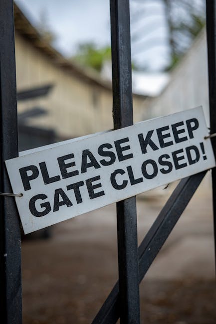 A close-up view of a rectangular 'No Trespassing' sign attached to a glass door or window. The sign features a black background with bold, red uppercase letters spelling 'NO TRESPASSING' at the top and a blank white rectangular space beneath for additional information. The sign is secured with multiple pieces of white tape around its edges, affixed to transparent glass, which reflects a faint outdoor scene showing residential buildings with multiple windows and a cloudy sky. The environment suggests an urban or suburban setting. The sign's presence indicates restricted access to the property, typically managed through private notices, aligning with alternative waste management or property access control measures commonly used by waste removal services such as Waste Removal Enfield that operate independently of local authority rubbish collection.