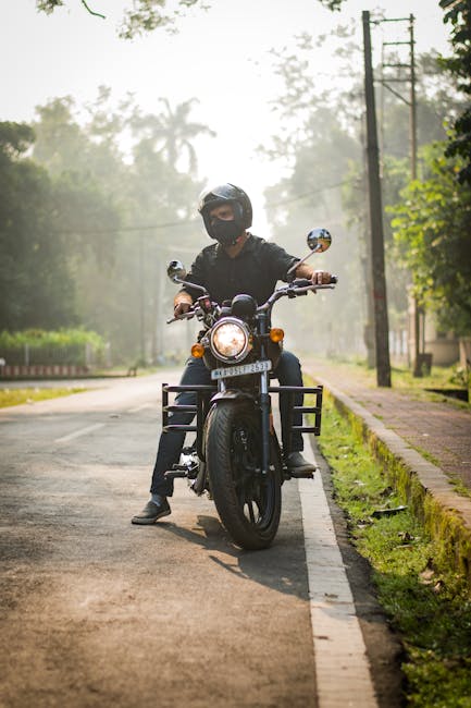 A person wearing a black helmet and face mask is seated on a black motorcycle positioned on a quiet residential street, facing directly towards the camera. The motorcycle has a large round headlight that is turned on, with chrome side mirrors and metal frames on either side of the front wheel. The individual is dressed in dark clothing, and their hands rest on the handlebars. The street features a paved road with a narrow white line along the edge, bordered by a grassy verge and a brick-paved footpath on the right side. In the background, there are tall wooden utility poles with power lines running parallel to the street, and lush green trees and shrubs providing a dense, leafy environment. The lighting suggests early morning or late afternoon, with a soft, diffused glow creating a calm atmosphere. This scene subtly relates to the context of private or alternative waste transportation methods, as the motorcycle could be used for quick, independent travel, similar to how some waste removal professionals might operate within local communities, exemplifying on-site or non-standard waste collection practices.