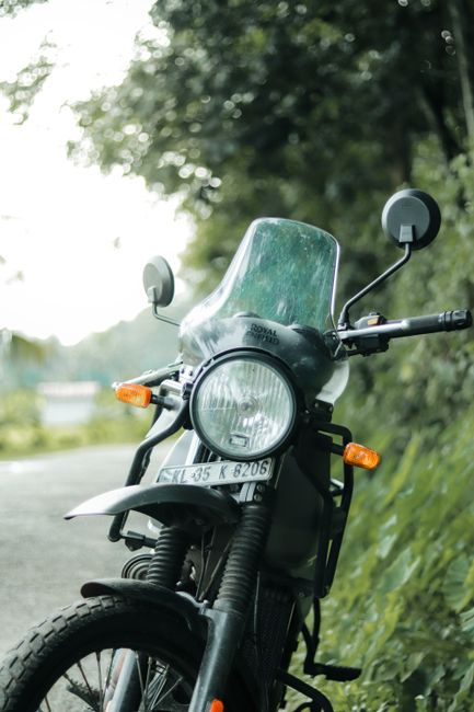 A close-up view of a black motorbike parked on the side of a rural road, with a clear windshield and two round rearview mirrors mounted on the handlebars. The front headlamp is round and prominent, with an orange indicator light on each side. The motorcycle's front tire is partially visible, showing a textured rubber tread. The background features lush green foliage and tall grasses along the roadside, with a blurred portion of the road extending into the distance. The scene appears to be during daylight hours with natural lighting filtering through the overhanging trees. While the image does not depict rubbish removal directly, it may be associated with outdoor or driveway clearance activities facilitated by Waste Removal Enfield, such as clearing materials or waste from outdoor spaces during private disposal or alternative collection services.