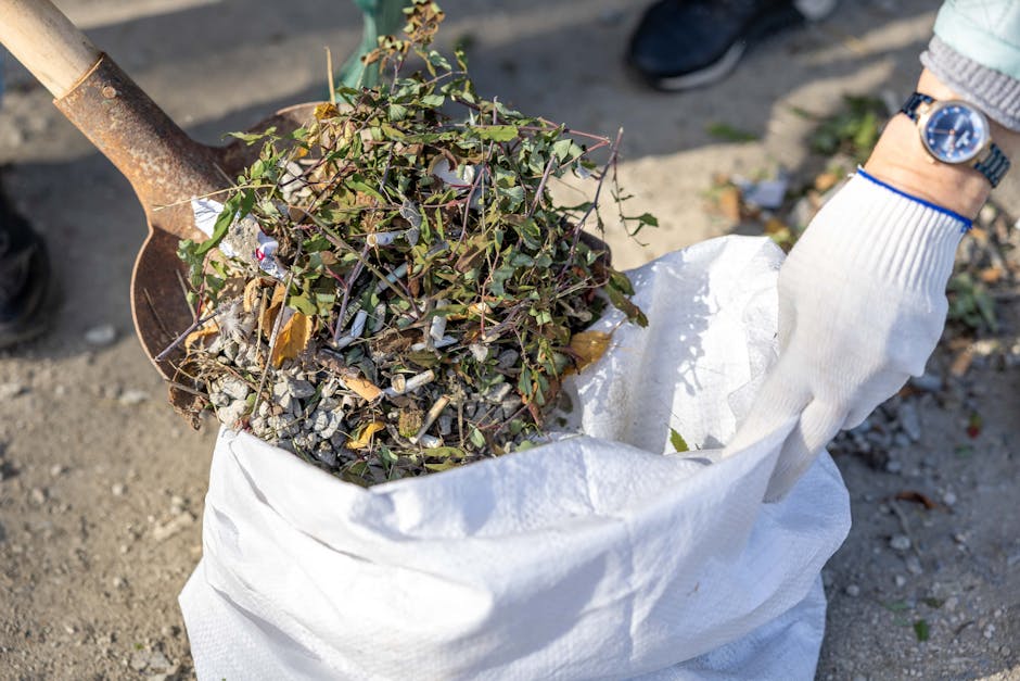 A person wearing a white work glove is holding a white trash bag filled with mixed garden debris, including small green leafy branches, twigs, and scattered gravel and stones. The debris appears to include natural organic matter alongside some small, light-colored rocks. The bag is partially open, revealing the contents, and is situated outdoors on a dirt or gravel surface. A garden spade with a wooden handle and metal blade is positioned inside the bag, its blade visible above the foliage. Nearby, a person is wearing dark shoes and a wristwatch, indicating participation in a yard clean-up or garden clearance activity. The background features natural outdoor elements and a focus on waste collection suitable for garden waste removal services, such as those offered by Waste Removal Enfield, supporting the context of independent garden debris disposal or alternative waste handling solutions.