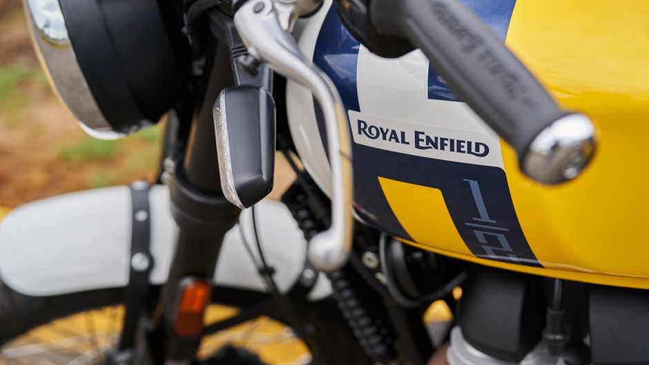 A close-up view of a black motorbike parked on the side of a rural road, with a clear windshield and two round rearview mirrors mounted on the handlebars. The front headlamp is round and prominent, with an orange indicator light on each side. The motorcycle's front tire is partially visible, showing a textured rubber tread. The background features lush green foliage and tall grasses along the roadside, with a blurred portion of the road extending into the distance. The scene appears to be during daylight hours with natural lighting filtering through the overhanging trees. While the image does not depict rubbish removal directly, it may be associated with outdoor or driveway clearance activities facilitated by Waste Removal Enfield, such as clearing materials or waste from outdoor spaces during private disposal or alternative collection services.