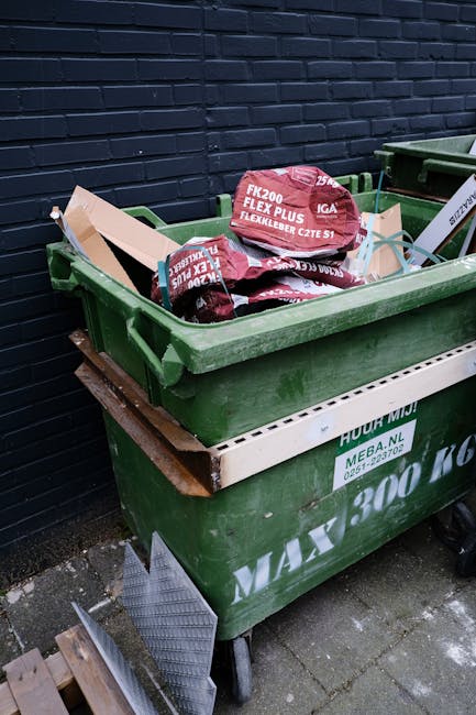 A large green waste bin positioned on a paved surface against a dark brick wall, filled with red and black bags labeled 'FK200 FLEXY PLUS' containing building or garden waste; a white plastic pallet or container is placed underneath the bin, and a small section of another green bin is visible to the right. The scene is outdoors, with natural lighting highlighting the textured surface of the brick wall and the contents of the bin, indicating an arrangement for private waste disposal or rubbish clearance by Waste Removal Enfield, reflecting an alternative approach to typical council rubbish collection.