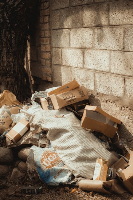 A collection of discarded cardboard boxes, some flattened and others partially inflated, along with crumpled paper and plastic packaging, piled against a brick wall outdoors. The boxes vary in size, with some showing printed labels and barcodes, and have a brown corrugated texture. A large, white woven sack or bag is partially buried within the pile, adding a fabric element to the scene. The rubbish is situated at the base of a wall made of concrete blocks with a weathered appearance, in front of a tree trunk with rough, dark bark. The environment appears to be an outdoor, urban or semi-urban setting, close to a building structure, with natural light illuminating the scene. The arrangement of waste suggests an outside private rubbish collection or disposal activity, potentially handled by local waste removal services such as Waste Removal Enfield, supporting alternative waste handling outside of regular municipal collections.