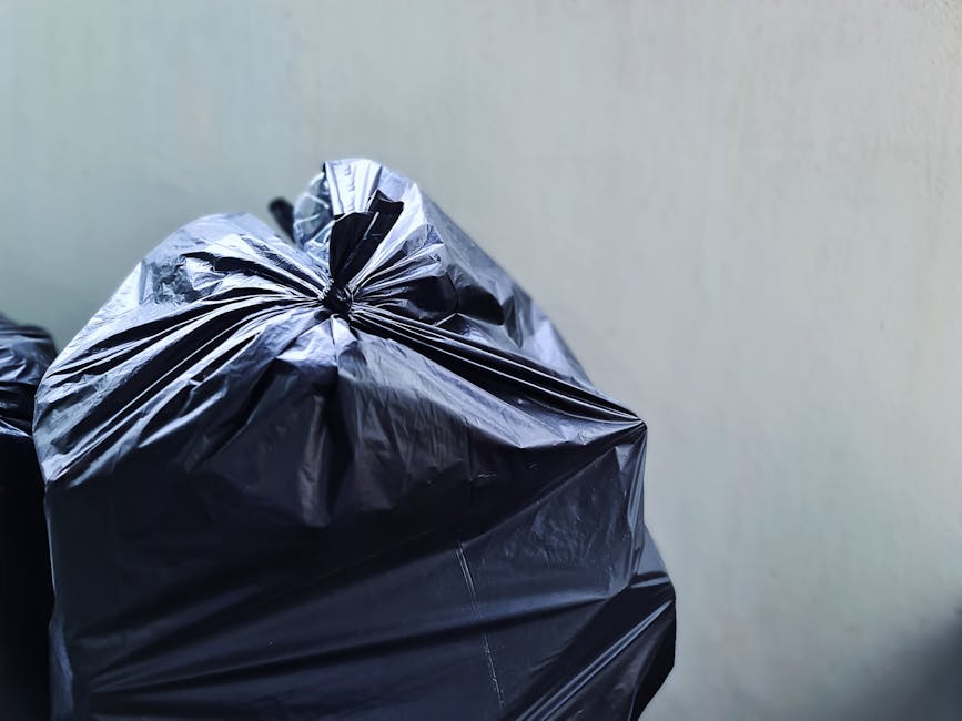 Close-up of a tightly tied black plastic rubbish bag with a shiny, crinkled surface, positioned in the left foreground against a plain, pale-colored wall. The bag appears full and is resting on a surface that is not visible in the image. In the background, additional black rubbish bags are visible, slightly out of focus, stacked or placed alongside the primary bag, suggesting a collection or disposal of waste materials. The environment appears to be an indoor or enclosed space, possibly a storage area or a designated rubbish deposit zone. The lighting is even and neutral, with no strong shadows or highlights, emphasizing the utilitarian nature of the scene. The image visually supports the context of private waste handling or rubbish collection, typical of services provided by companies like Waste Removal Enfield, which specialise in alternative waste disposal options outside of municipal collection services.