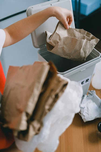 A person’s hand is shown placing a crumpled brown paper bag into a small, white, open-top rubbish bin with a black interior, situated on a light-colored surface in an indoor setting. The bin, labeled with partially visible black text, is positioned near a wooden table or counter. In the foreground, a stack of additional crumpled brown paper bags is held, ready to be disposed of. The background includes blurred objects and a sleek, modern environment, possibly an office or kitchen area. The scene depicts a routine act of waste disposal, highlighting a conscious approach to eco-friendly rubbish handling, which is associated with private waste collection or on-site rubbish removal services. The lighting is soft, natural or artificial, providing a clear view of the materials and textures: the coarse, slightly wrinkled surface of the paper bags, the smooth, clean finish of the rubbish bin, and the contrasting tones of the brown paper against the white container. This image emphasizes individual waste management practices suitable for professional rubbish removal contexts.