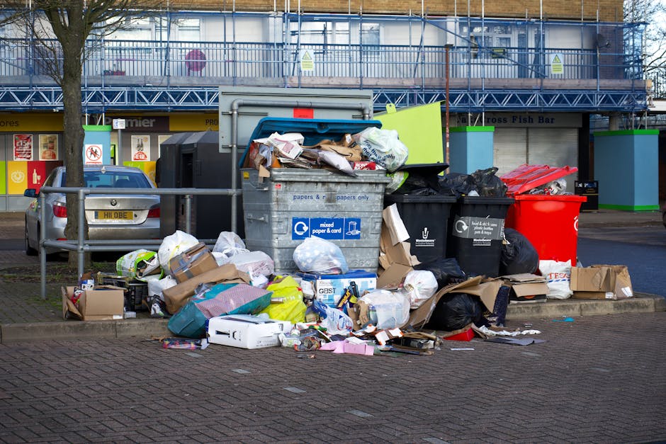 A large outdoor rubbish collection point situated on a paved area in front of a commercial building with a blue metal framework and signage, featuring multiple waste containers overflowing with mixed household and office waste. The central gray bin labeled for mixed paper and card is open, with various flattened cardboard boxes, crumpled paper, and plastic packaging spilling onto the ground. Surrounding it are black and red waste bags, some tied and others open, revealing additional refuse inside, along with loose paper, packaging, and smaller discarded items scattered across the pavement. A silver car is parked nearby on the left side, partially obscured by a metal railing, which separates the waste area from the parking space. The scene is illuminated by natural daylight, with the background showing parts of the building’s shops or services and a leafless tree on the left, indicating the scene takes place outdoors in an urban area, typical of private waste handling or independent collection points managed by waste removal services like Waste Removal Enfield.