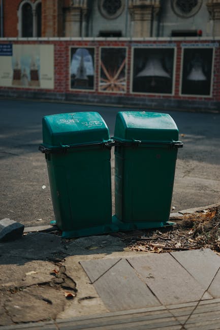 Two green wheeled rubbish bins with hinged lids and black handles are positioned side by side on a paved sidewalk near the edge of a road. The bins are made of plastic with a smooth, slightly reflective surface, and the lids are closed, showing some minor scratches and scuff marks. They are placed on a gray stone or concrete base, which is adjacent to a section of uneven pavement with patches of dirt, fallen leaves, and small debris visible around the bases. In the background, there is a street with a dark asphalt surface and painted road markings, and behind it an old brick building featuring decorative arched windows, some with white trim, and a series of posters or advertisements mounted on the wall. The scene appears to be in daylight with ambient natural light, illustrating a typical urban setting where private rubbish disposal might be arranged through services like Waste Removal Enfield, supporting alternative waste handling options outside of local authority collection.