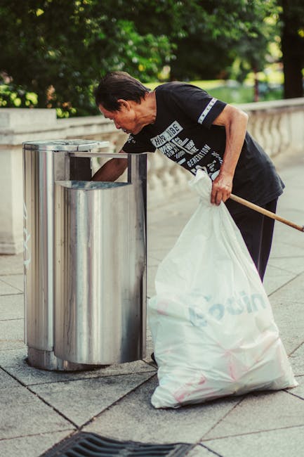 A middle-aged man with dark hair is seen standing on a paved urban walkway, emptying a large white waste bag into a metallic rubbish bin with a rounded, polished surface. The man is wearing a black T-shirt with white printed text and a dark pair of trousers. He holds a wooden stick in his right hand, which he is using to assist in guiding the bag into the bin. The metallic rubbish bin is positioned near a low stone wall with rounded edges, and behind it, there are lush green trees and foliage, indicating a park or landscaped area. The sunlight filters through the leaves, casting gentle shadows on the pavement, creating a bright and clean environment focused on waste disposal. This scene demonstrates independent waste handling, reflecting the importance of local rubbish collection and on-site clearance services, such as those offered by Waste Removal Enfield, capturing a practical aspect of rubbish management in urban settings.