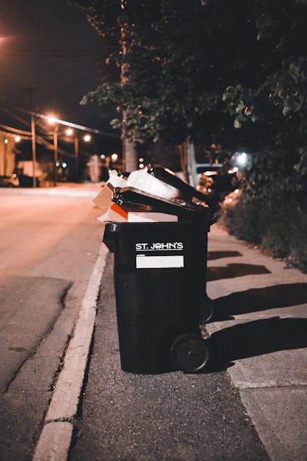A middle-aged man with dark hair is seen standing on a paved urban walkway, emptying a large white waste bag into a metallic rubbish bin with a rounded, polished surface. The man is wearing a black T-shirt with white printed text and a dark pair of trousers. He holds a wooden stick in his right hand, which he is using to assist in guiding the bag into the bin. The metallic rubbish bin is positioned near a low stone wall with rounded edges, and behind it, there are lush green trees and foliage, indicating a park or landscaped area. The sunlight filters through the leaves, casting gentle shadows on the pavement, creating a bright and clean environment focused on waste disposal. This scene demonstrates independent waste handling, reflecting the importance of local rubbish collection and on-site clearance services, such as those offered by Waste Removal Enfield, capturing a practical aspect of rubbish management in urban settings.